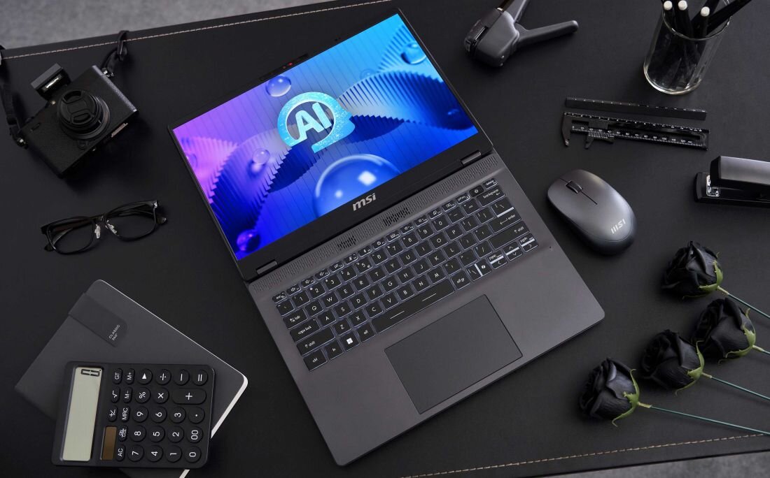 Laptop MSI Prestige 14 AI Studio na czarnym biurku w otoczeniu akcesoriów biurowych, aparat fotograficzny, czarne róże, kalkulator i okulary, ekran z grafiką AI, podświetlana klawiatura.