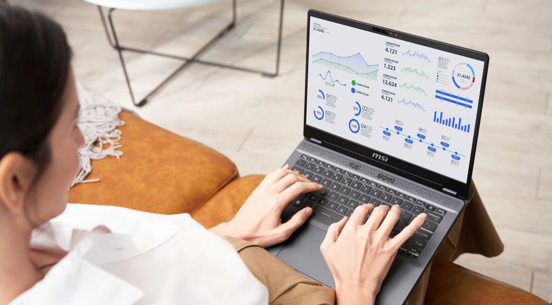 Osoba pracująca na laptopie MSI Prestige 14 AI Studio, wyświetlającym wykresy i dane analityczne na ekranie, siedzi wygodnie na brązowej sofie, jasne wnętrze i stół w tle. - RAM DDR5 5600 MHz, Dysk SSD PCIe NVMe 4.0