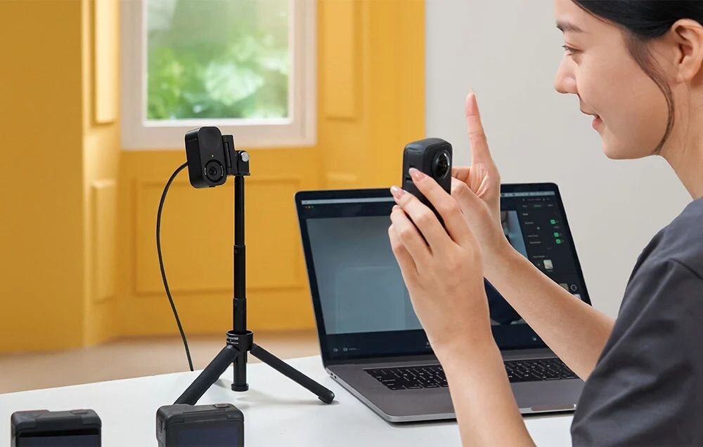 insta360 Link 2C insta360 Link 2C rozmyć tło zastąpić je wybranym obrazem  ustawienia i filtry obrazu One-Click Makeup  - Kobieta przy laptopie trzymająca w dłoni niewielką kamerę obok drugiej kamery na małym statywie..