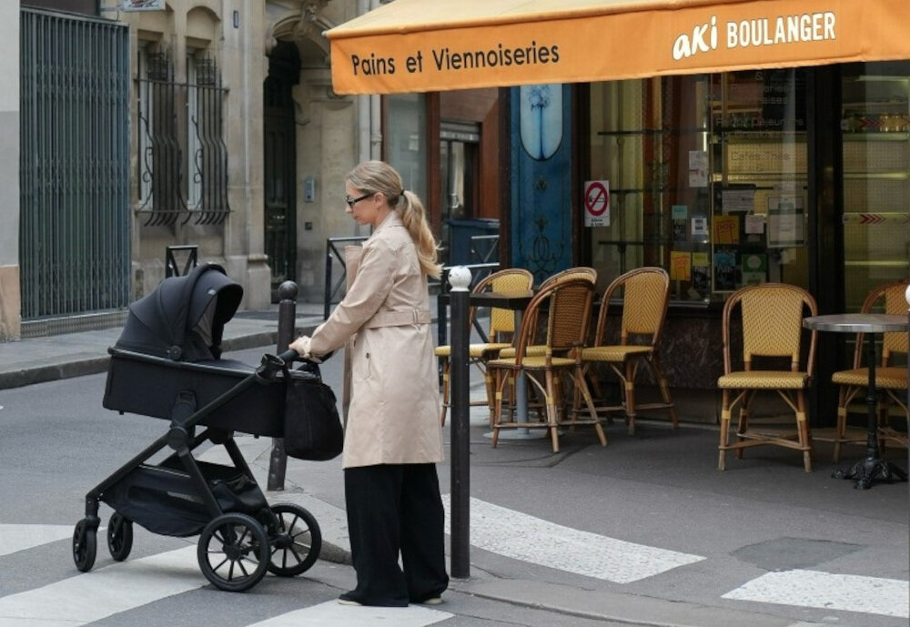 Osoba stoi obok czarnego wózka dziecięcego na ulicy, przed kawiarnią z żółtym daszkiem z napisem \'Pains et Viennoiseries aki BOULANGER\'.