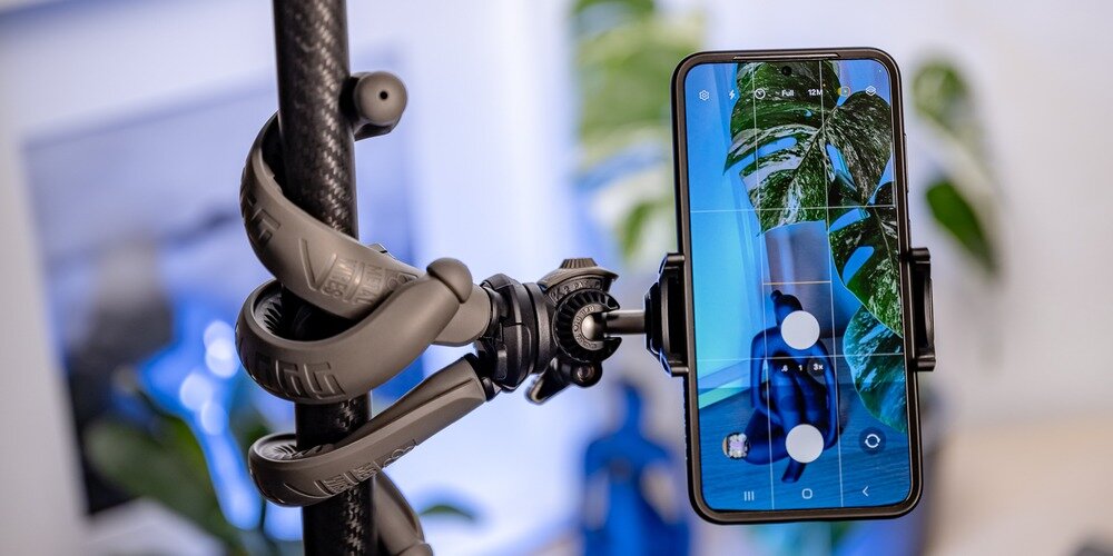 Smartfon zamocowany na elastycznym statywie Newell, owiniętym wokół rurki, z podglądem kadru rośliny na ekranie
