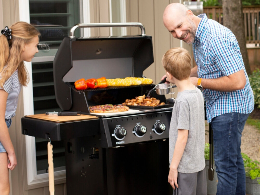 Grill gazowy BROIL KING Crown 410 Shadow 11.4 kWh 65 x 44 - 64 x 17 cm trzy osoby stoją przy grillu śmieją się grillują potrawy w tle dom grillowanie czas na świeżym powietrzy przyrządzanie potraw pieczenie mięsa i warzyw