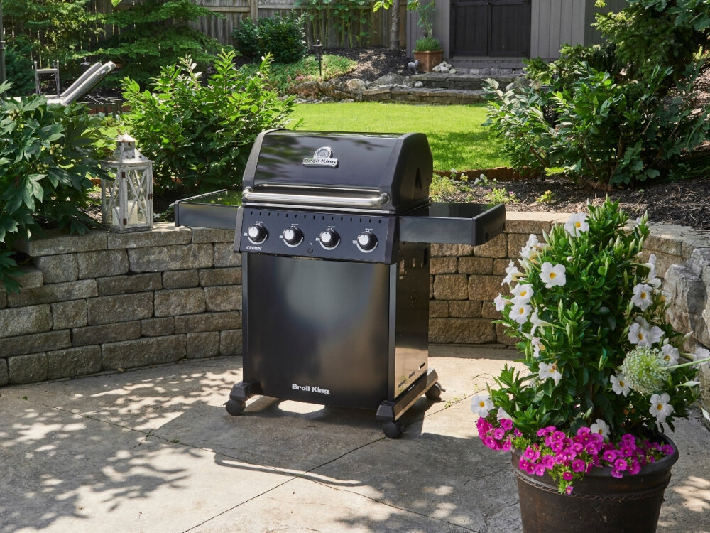 Grill gazowy BROIL KING Crown 410 Shadow 11.4 kWh 65 x 44 - 64 x 17 cm ogróg grill ustawiony na tarasie w tle rośliny i trawnik eleganckie czarne wykończenie półki boczne drzwi panel kontrolny pomalowane czarną farbą proszkową zaawansowane technologie