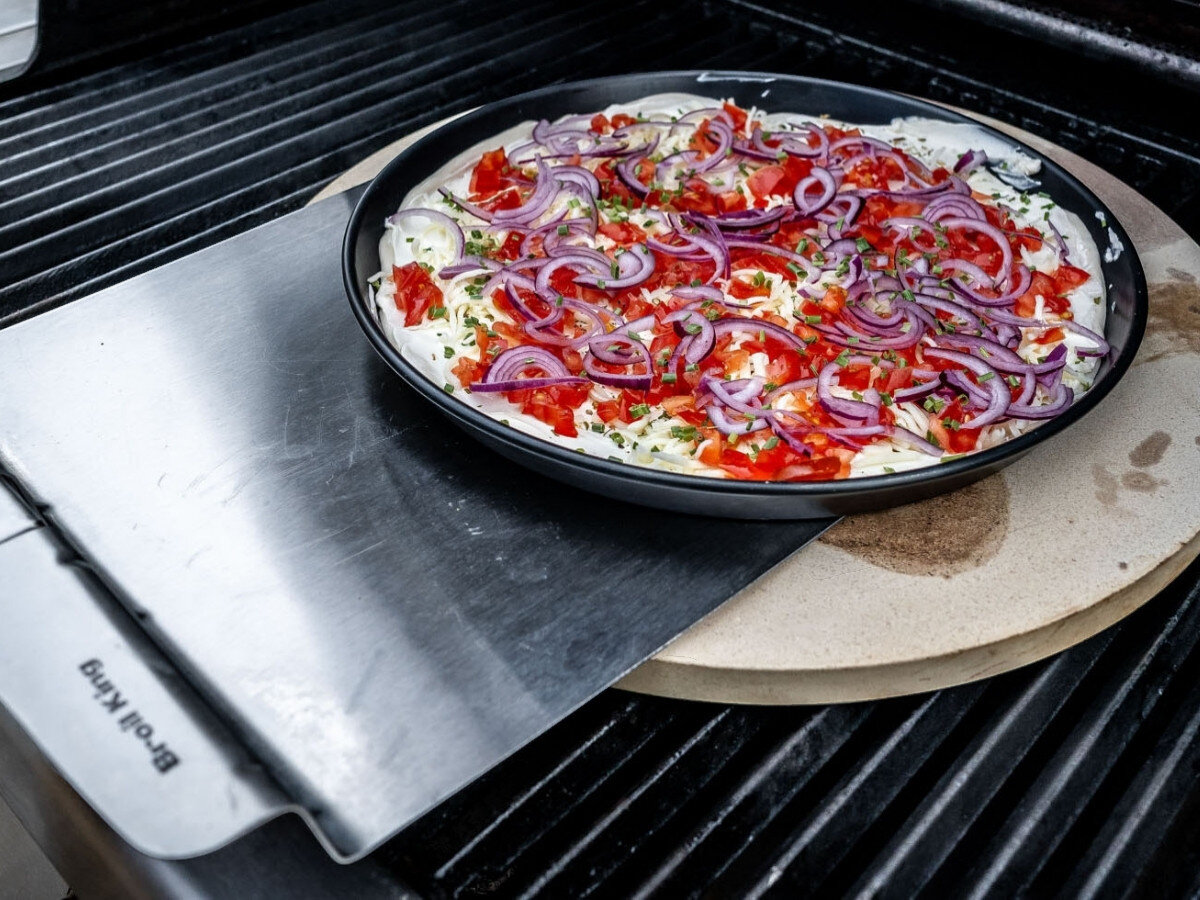 Kamień do pizzy BROIL KING 33 cm, kamień, focaccia, pieczywo, tarty, Ręce w rękawicach wyjmują pizzę z mozzarellą, szparagami, grzybami i artichoke z grilla na drewnianej desce.