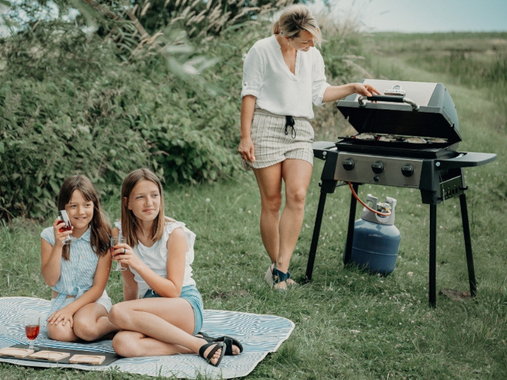 Grill gazowy BROIL KING Porta-Chef 320 6 kW 53 x 34 - 46.5 x 18.5 cm łąka jedna osoba stoi przy grillu obok na kocu siedzą dwie osoby grillowanie czas na świeżym powietrzy przyrządzanie potraw pieczenie mięsa i warzyw