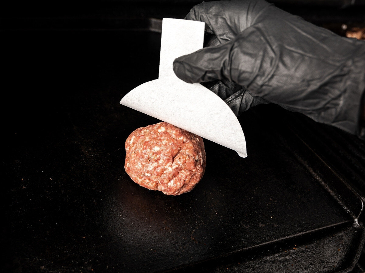 Papier do praski BROIL KING 62470 (100 szt.), formowanie, papier, grill, ułatwienie, Ręka w czarnej rękawicy kładzie burgera na białej papierowej szpachelce na płycie grilla, z zaznaczoną rączką.