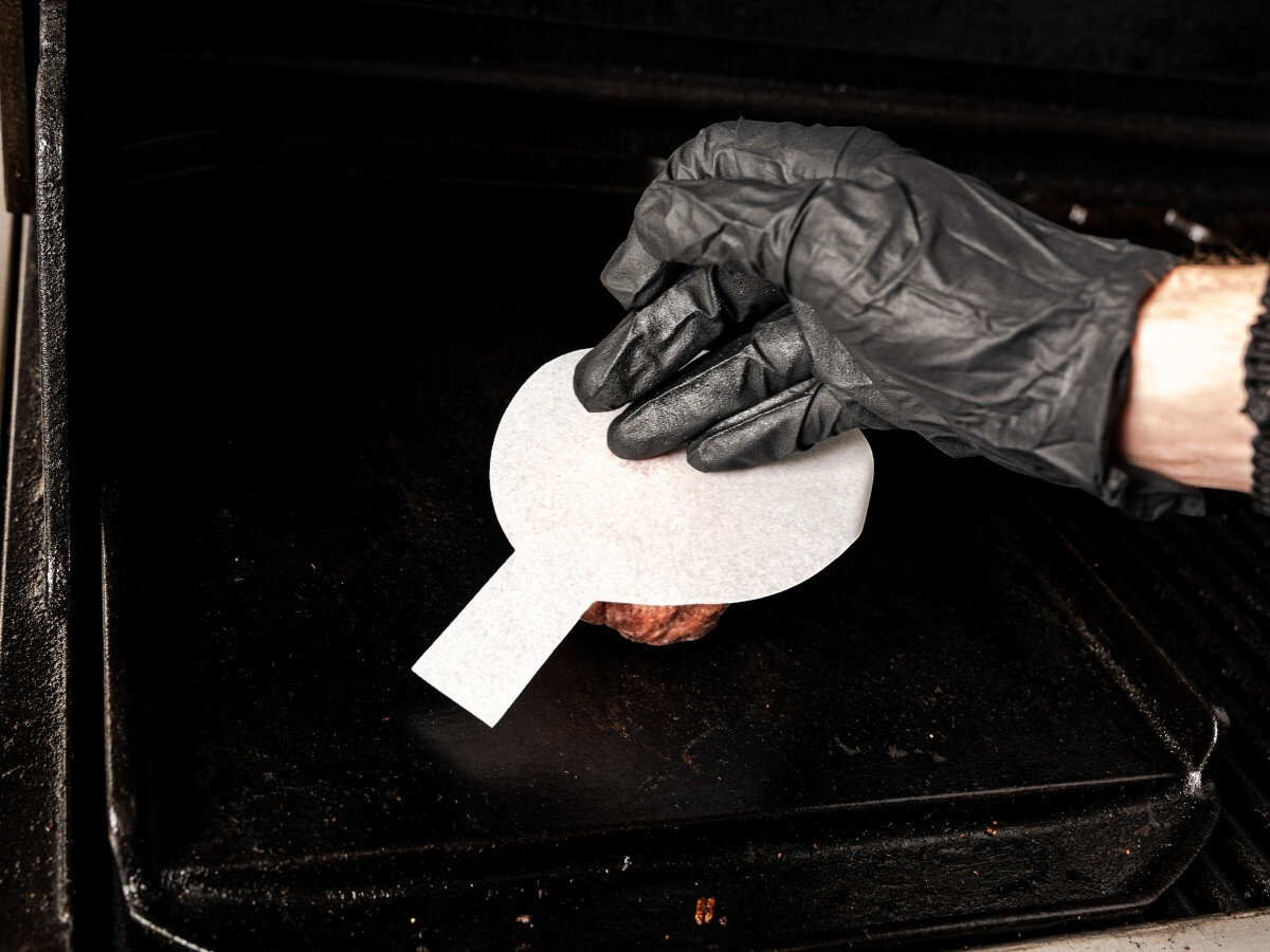 Papier do praski BROIL KING 62470 (100 szt.), papier, średnica, małe, duże, Ręka w czarnej rękawicy dociska białą papierową szpachelkę do smażenia burgera na czarnej płycie żeliwnej grilla.