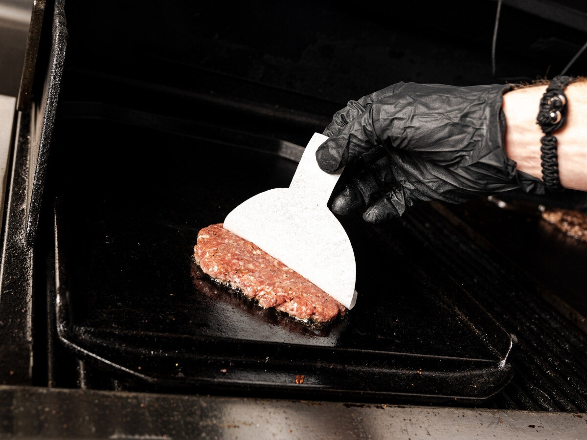 Papier do praski BROIL KING 62470 (100 szt.), pasek, chwytanie, papier, wygoda, Ręka w rękawicy smaruje burgera na płycie grilla za pomocą białej papierowej szpachelki z wystającą rączką.