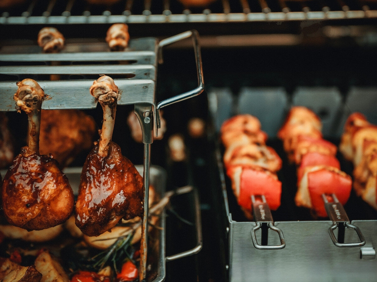 Wieszak na podudzia BROIL KING 64152, stal, zmywarka, trwałość, czyszczenie, Bliski widok uchwytu rotacyjnego Broil King z udkami kurczaka nad blaszką z warzywami i pieczarkami obok grilla z szaszłykami.