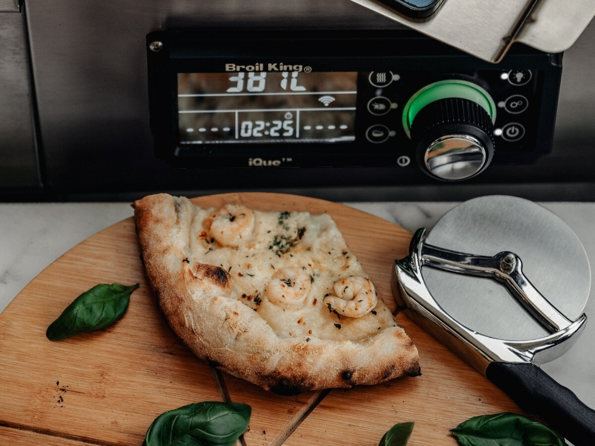 Nóż do pizzy BROIL KING 69810, stal, trwałość, ostrze, szlachetna, Kawałek pizzy ze śmietaną i krewetkami na drewnianej desce obok panelu IQ piekarnika Broil King pokazującego czas pieczenia 02:25 i temperaturę.