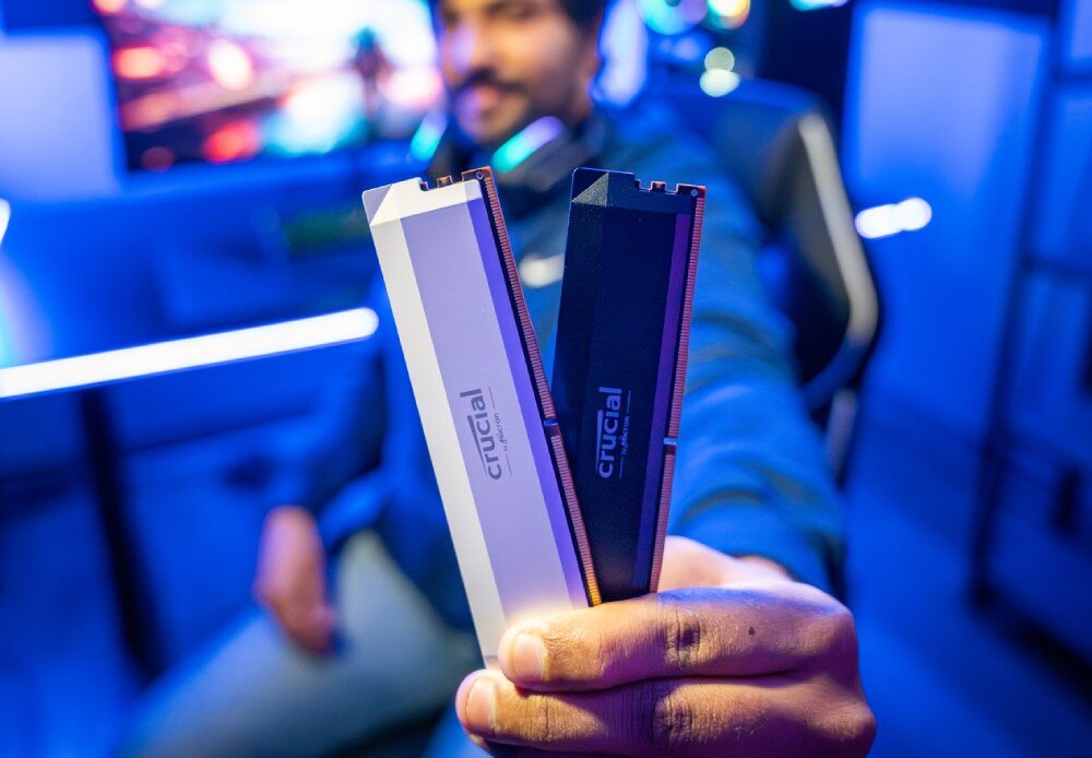 Pamięć RAM CRUCIAL 16GB 6000MHz CP16G60C36U5B Osoba trzymająca w dłoni dwa moduły pamięci, jeden czarny i jeden biały, z rozmytym tłem stanowiska gamingowego, typ pamięci, taktowanie, opóźnienia, wydajność