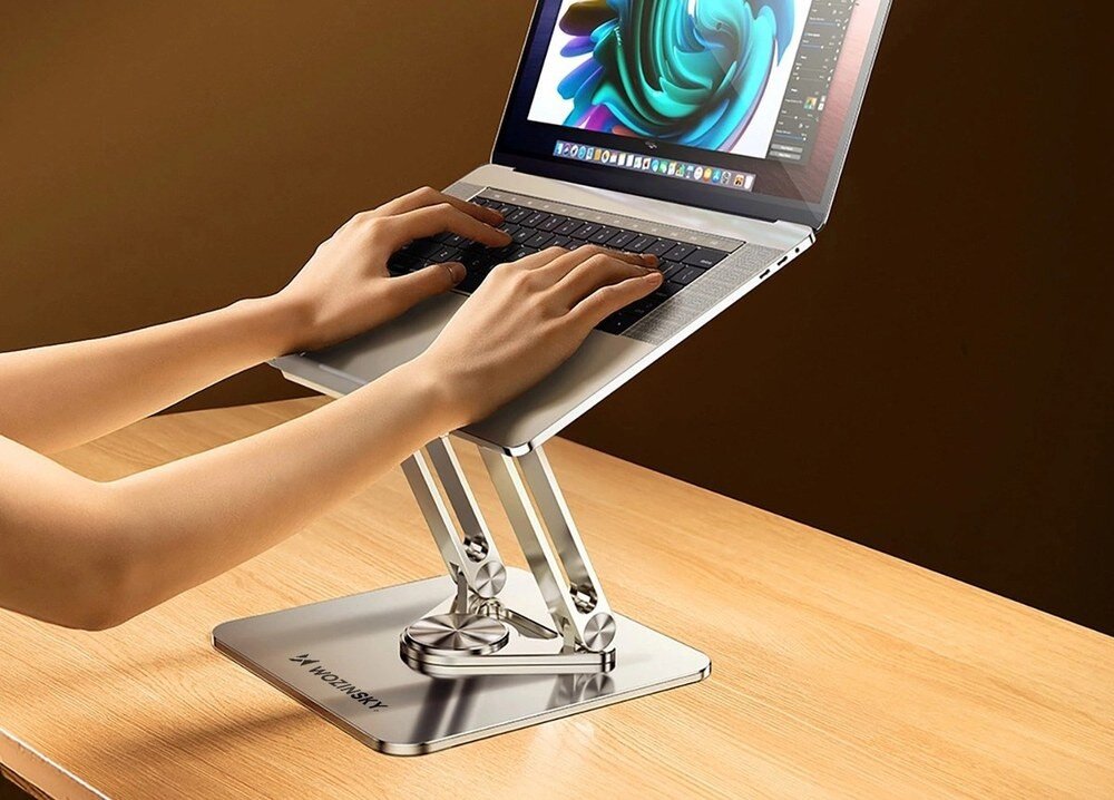 Srebrna podstawka pod laptopa z logo \'WOZINSKY\' podtrzymuje otwarty laptop na drewnianym biurku. Na ekranie laptopa widoczna jest kolorowa grafika, a ręce osoby piszą na klawiaturze.