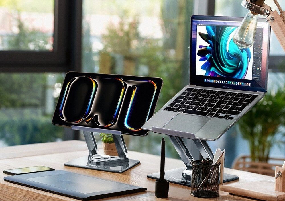 Na biurku znajdują się dwa urządzenia elektroniczne umieszczone na srebrnych podstawkach, jedno z nich to laptop z otwartym ekranem. W tle widoczne jest rozmyte wnętrze pomieszczenia z dużymi oknami.