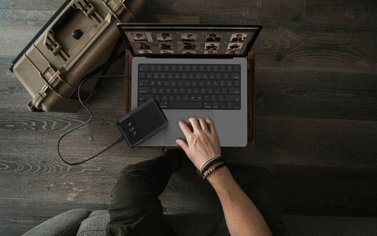Laptop na drewnianym biurku podpięty do power banku BELKIN, widoczna jest dłoń dotykająca touch pad laptopa.