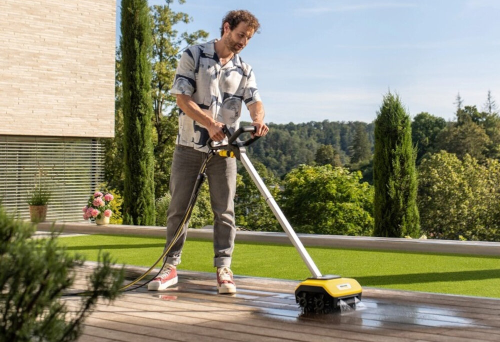 Osoba używa urządzenia do czyszczenia posadzek na drewnianym tarasie. W tle widoczna jest zieleń i nowoczesny budynek.