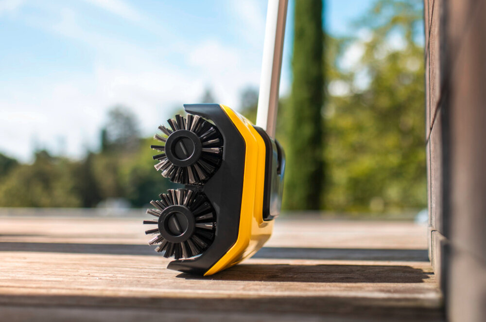 Urządzenie do czyszczenia posadzek z dwoma okrągłymi szczotkami, w żółtej obudowie, znajduje się na drewnianej powierzchni. W tle widoczna jest rozmyta zieleń i niebo.