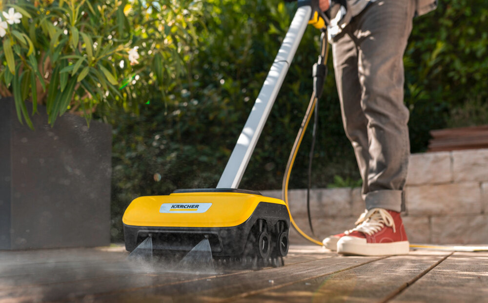 Osoba używa urządzenia do czyszczenia posadzek z logo KARCHER na drewnianej powierzchni. W tle widoczne są rośliny i kamienny mur.