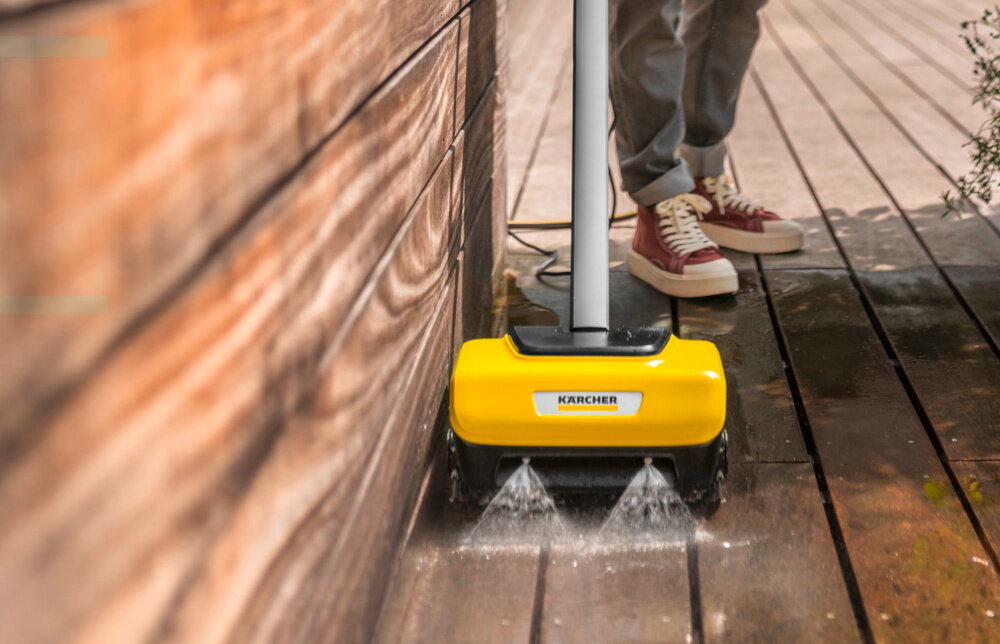 Urządzenie do czyszczenia posadzek KARCHER w kolorze żółtym, z widocznym logo \'KARCHER\', używane na drewnianej powierzchni. W tle widoczne są nogi osoby w czerwonych butach.