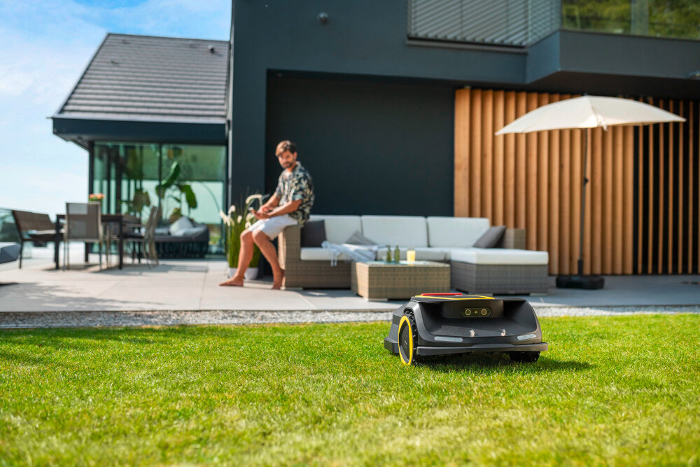 Robot koszący KARCHER RCX 6 1.269-760.0 sterowanie Wi-Fi-Bluetooth robot na trawie w tle osoba siedzi na oparciu narożnika ogrodowego z tyłu dom aplikacja Outdoor Robots z dostępem do harmonogramu stref i wysokości cięcia wyświetlacz LCD blokada PIN zabezpieczenie antykradzieżowe bezpieczeństwo aktualizacje oprogramowania OTA
