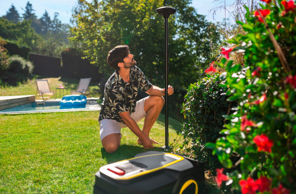 Osoba klęczy na trawie obok robota koszącego, trzymając pionowy pręt. W tle widoczny jest basen oraz krzewy z czerwonymi kwiatami.