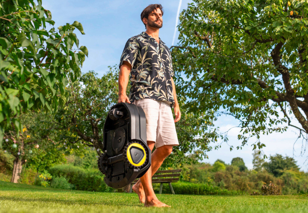 Mężczyzna w koszuli z wzorem roślinnym i krótkich spodenkach trzyma w ręku czarny robot koszący z żółtym akcentem. W tle widać zielony ogród z drzewami i ławką.
