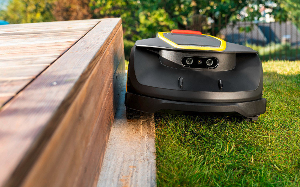 Robot koszący porusza się wzdłuż drewnianej krawędzi na trawniku. Urządzenie ma czarną obudowę z żółtymi i czerwonymi akcentami.
