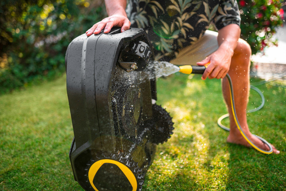 Osoba myje czarny robot koszący wodą z węża ogrodowego na trawniku. Robot ma żółte akcenty na kołach.