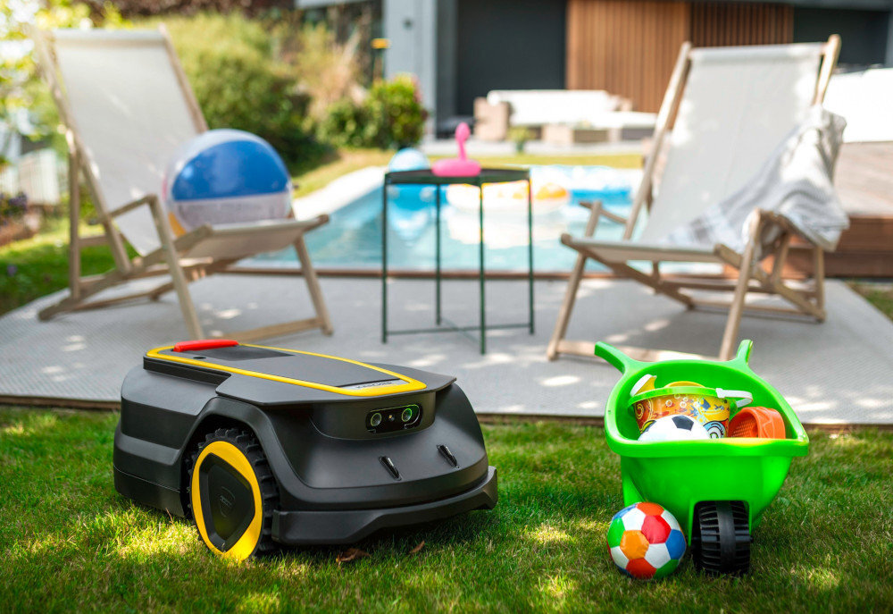 Na trawniku znajduje się robot koszący o czarno-żółtej kolorystyce. W tle widoczny jest basen, dwa leżaki oraz zielona taczka z zabawkami.