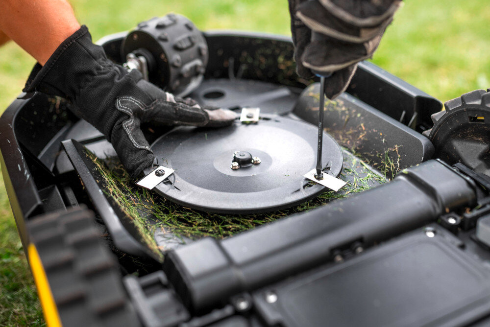 Osoba w rękawicach naprawia lub konserwuje spód robota koszącego, używając śrubokręta. Widać trawę przyczepioną do elementów urządzenia.