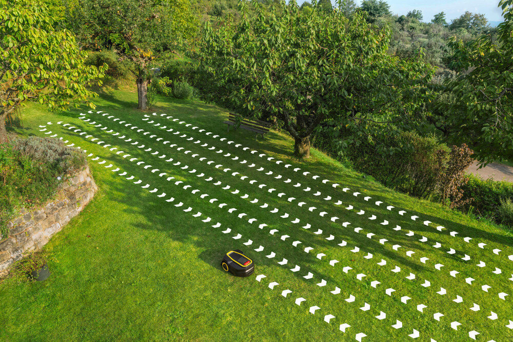Na trawniku w ogrodzie znajduje się robot koszący, a na trawie widoczne są białe strzałki wskazujące kierunek jego ruchu. W tle rosną drzewa i krzewy.