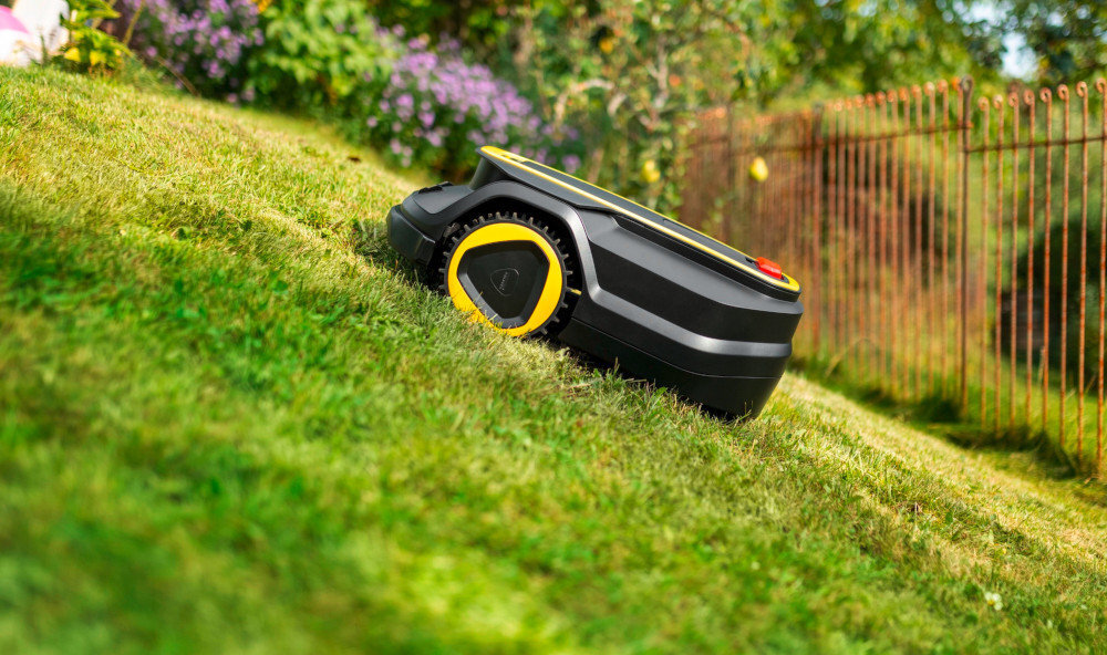 Robot koszący porusza się po trawniku na pochyłym terenie. W tle widoczne są kwiaty i metalowe ogrodzenie.