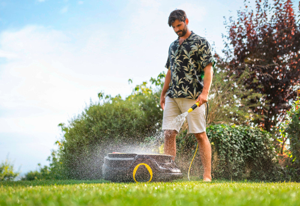 Osoba podlewa wodą z węża ogrodowego robot koszący na trawniku. W tle widoczne są krzewy i drzewa.