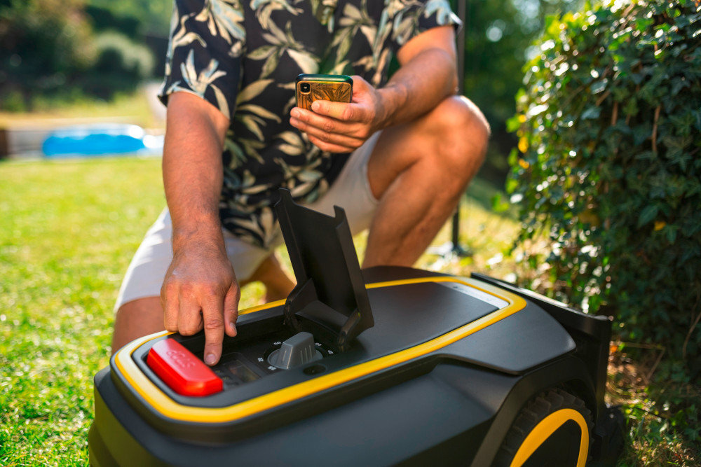 Osoba obsługuje robota koszącego, trzymając w ręku smartfon. Robot ma czarną obudowę z żółtymi akcentami i otwarty panel sterowania.