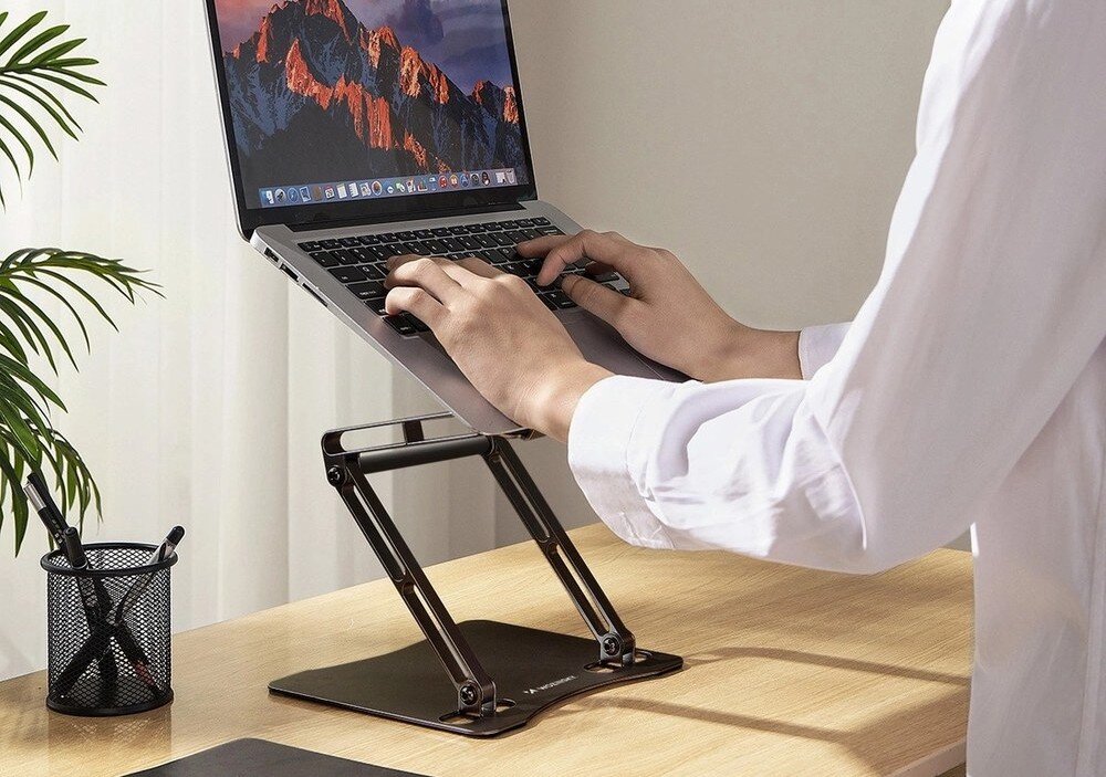 Osoba używa laptopa umieszczonego na czarnej podstawce na biurku. W tle widać zasłonę i doniczkę z rośliną, a obok laptopa stoi pojemnik z długopisami.
