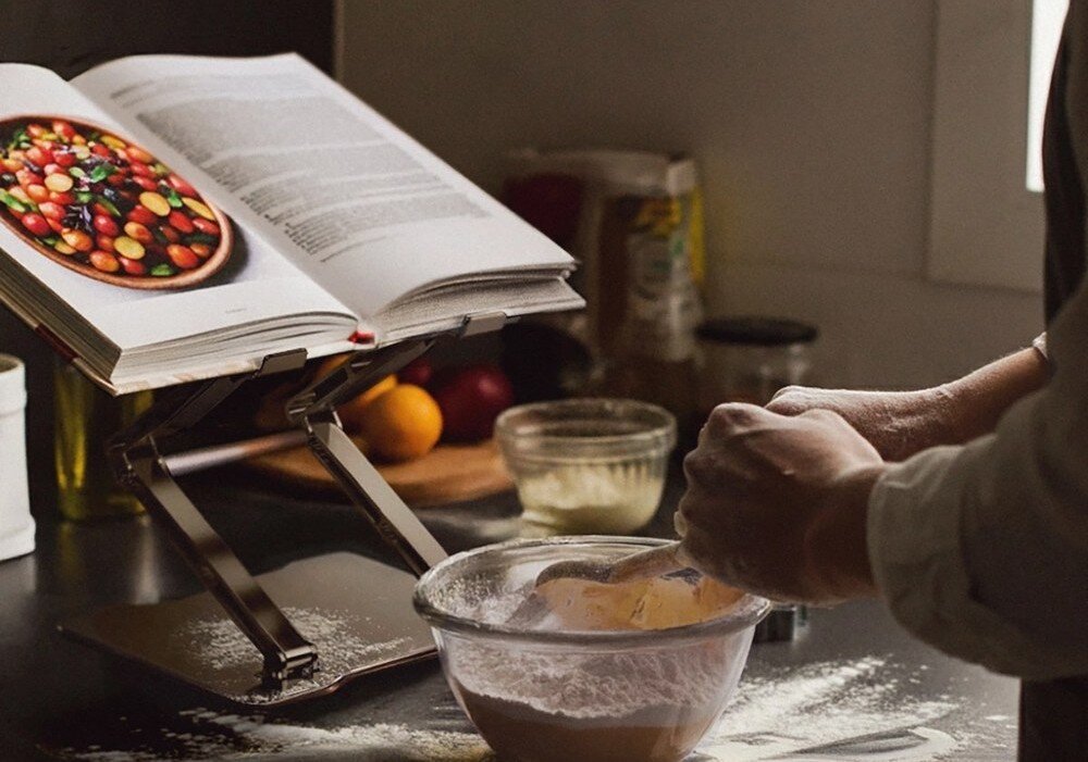 Na blacie kuchennym znajduje się otwarta książka kucharska na podstawce oraz miska z ciastem. W tle widoczne są różne składniki i przybory kuchenne.