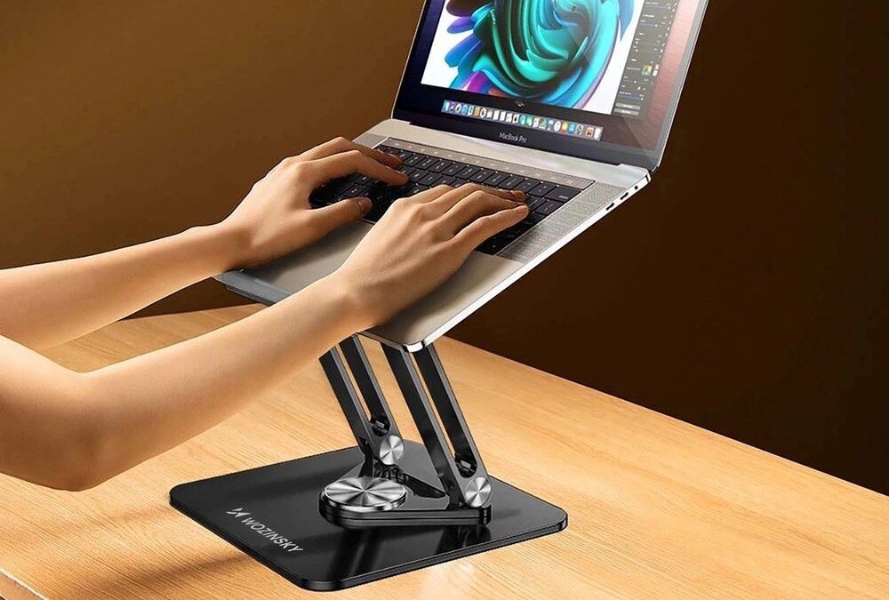 Na drewnianym biurku znajduje się czarna podstawka pod laptopa z logo \'WOZINSKY\', na której umieszczony jest otwarty laptop. Osoba pisze na klawiaturze laptopa, a na ekranie widoczny jest kolorowy wzór.