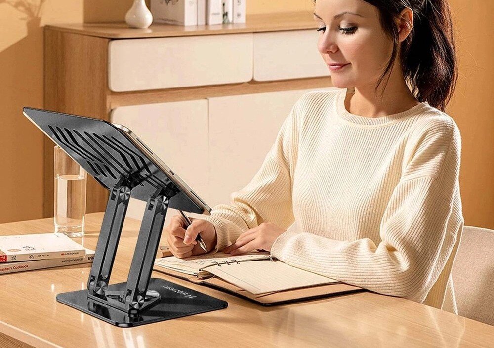 Na biurku znajduje się czarna podstawka pod laptopa z logo \'WOZINSKY\', na której umieszczony jest tablet. Obok siedzi osoba pisząca w notatniku.