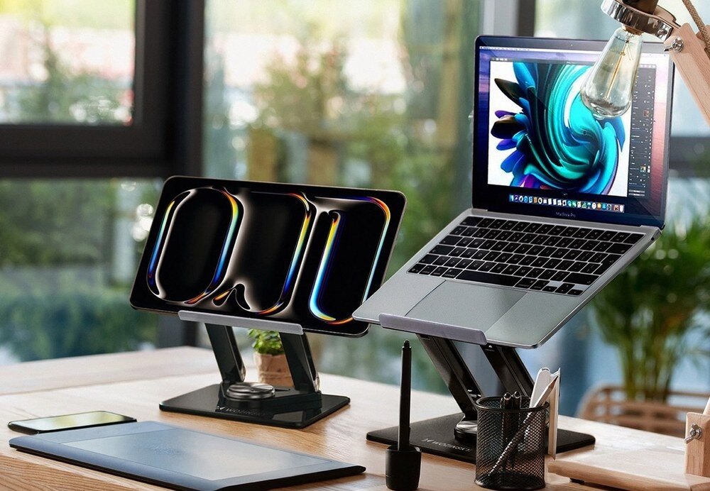 Na biurku znajdują się dwa urządzenia elektroniczne na czarnych podstawkach, jedno z nich to laptop z wyświetlonym kolorowym wzorem na ekranie. Obok widoczna jest lampka biurkowa oraz kilka akcesoriów biurowych.