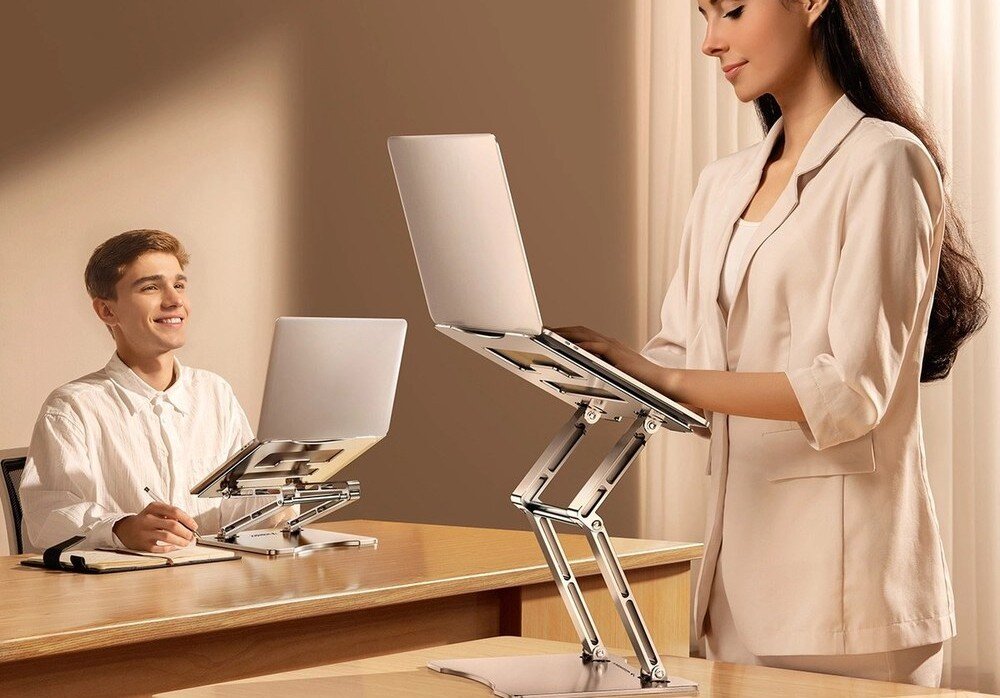 Dwie osoby korzystają z laptopów umieszczonych na srebrnych podstawkach na biurku. Podstawki mają regulowane ramiona, które podnoszą laptopy na wysokość oczu.