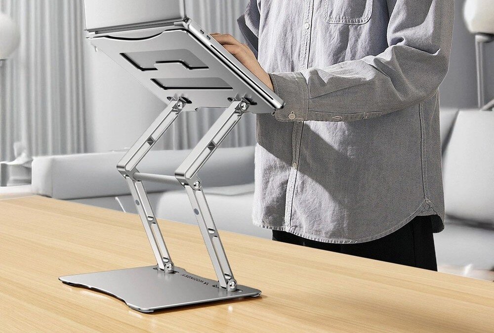 Osoba stoi przy biurku, używając srebrnej podstawki pod laptopa z regulowanymi ramionami. Na podstawce widoczne jest logo \'WOZINSKY\'.