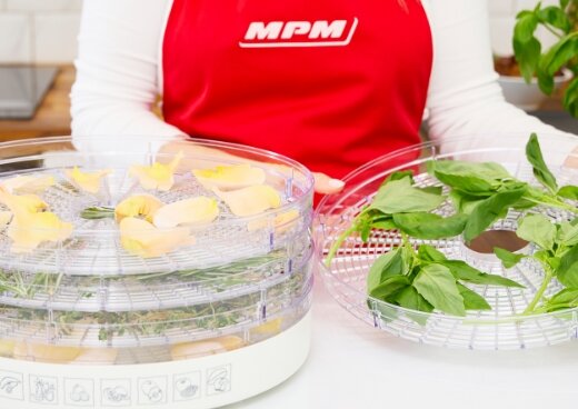 Osoba trzyma sito suszarki z ziołami. Obok suszarka z 4 sitami.