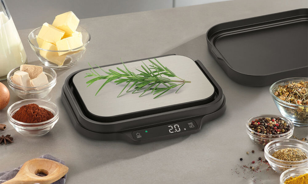 Waga kuchenna CASO KitchenDuo waga stoi na blacie wyświetlacz przyciski czarna podstawa srebrna góra na wadze zioła obok w miseczkach różne składniki z wysokiej jakości stali nierdzewnej trwałość odporność na zabrudzenia Czytelny wyświetlacz LED dotykowe sterowanie Funkcja HOLD zachowanie ostatniego pomiaru funkcja tary dokładne ważenie składników