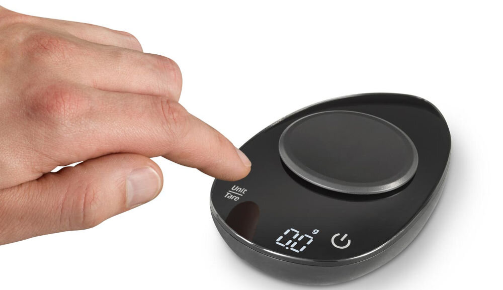Waga CASO FinoCompact osoba trzyma palec nad przyciskiem znajdującym się na wadze w kolorze czarnym białe tło zasilana dwiema bateriami AAA brak w zestawie mobilne urządzenie do pracy na wyjazd do laboratorium niski pobór energii precyzja wygoda stylowy design 