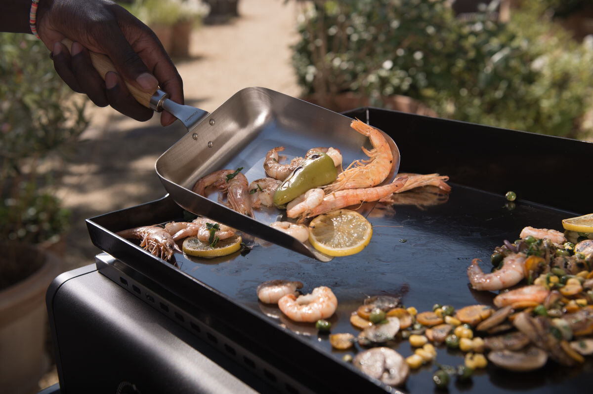 Grill gazowy CAMPINGAZ Plancha Othello 2, ścianki, boczne, wysokość, emaliowane, osoba przyrządza posiłek na grillu, w tle ogród
