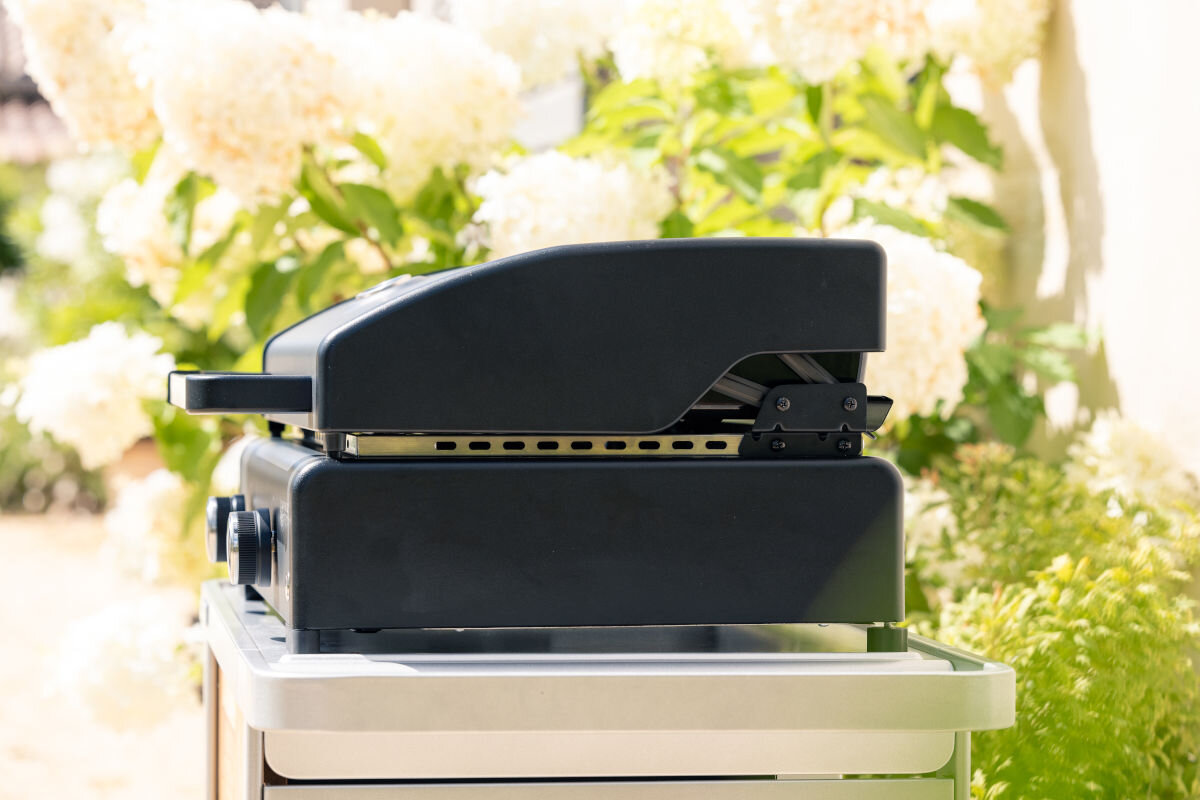 Grill gazowy CAMPINGAZ Plancha Sigma 2 cook, regulowane, nogi, płyta, wypoziomowanie, w tle ogród, na pierwszym planie grill od bocznej strony