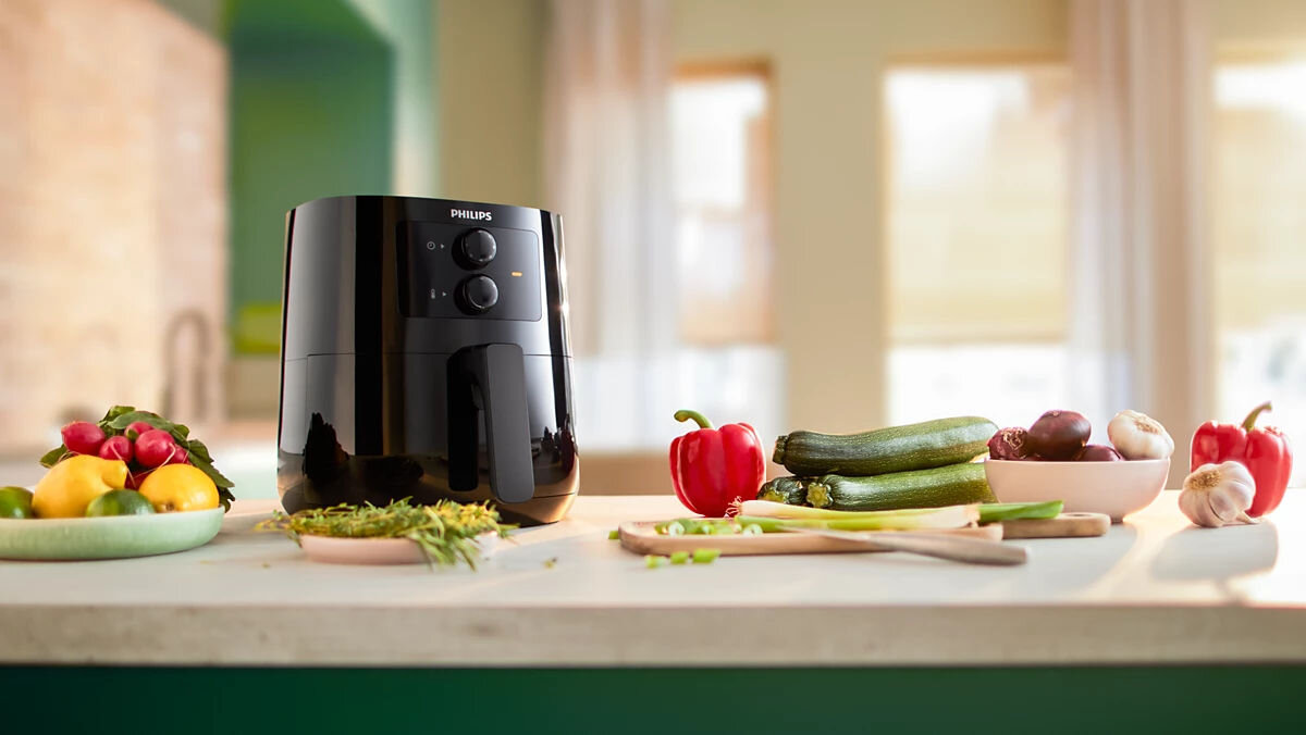 Air Fryer Frytkownica beztłuszczowa PHILIPS Ovi Mini Seria 3000 HD9200/90 stojąca na jasnym blacie kuchennym obok świeżych warzyw: papryki, cukinii, cebuli, czosnku i ziół zdrowy posiłek przygotowywany z użyciem frytkownicy beztłuszczowej Philips i świeżych warzyw