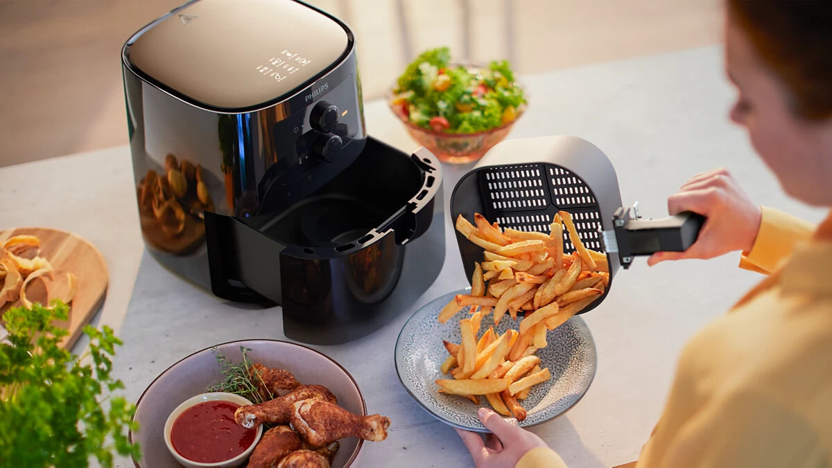 Air Fryer Frytkownica beztłuszczowa PHILIPS Ovi Mini Seria 3000 HD9200/90 osoba w jasnej koszuli wysypuje gorące, złociste frytki z kosza frytkownicy beztłuszczowej na talerz, obok widać miseczkę z sosem i usmażone udka kurczaka