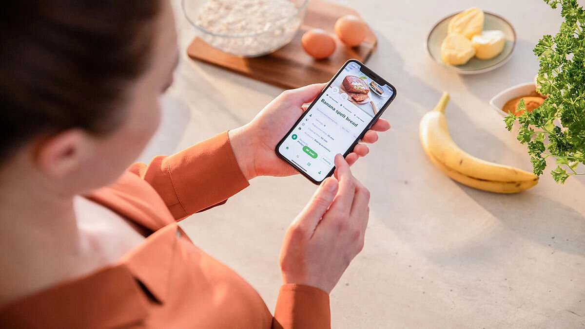 Air Fryer Frytkownica beztłuszczowa PHILIPS Ovi Mini Seria 3000 HD9200/90 Zbliżenie na dłonie kobiety trzymającej telefon z aplikacją kulinarną, na ekranie widnieje przepis na chleb bananowy orkiszowy, a na blacie obok przygotowane są składniki