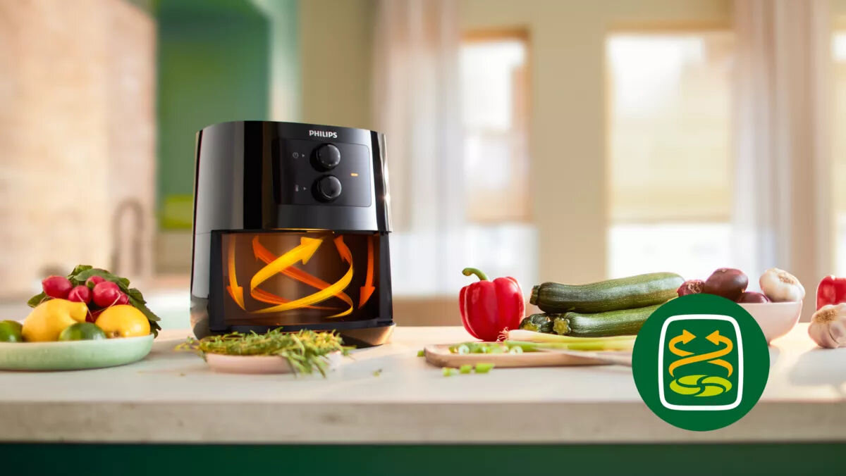 Air Fryer Frytkownica beztłuszczowa PHILIPS Ovi Mini Seria 3000 HD9200/90 Na blacie kuchennym stoi frytkownica beztłuszczowa marki Philips, obok niej znajdują się różnorodne warzywa, takie jak papryka, cukinia, rzodkiewki i czosnek, a także zielone zioła w prawym dolnym rogu widnieje zielone logo z dwoma strzałkami