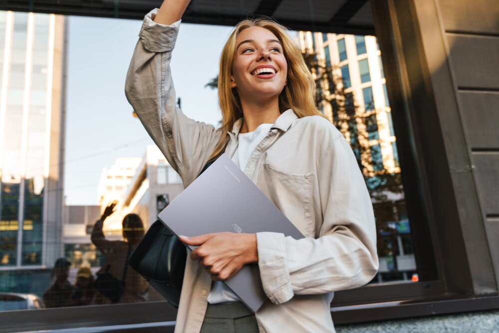 Laptop ASUS Chromebook Plus CX3402CVA Młoda kobieta w jasnej koszuli uśmiecha się i podnosi rękę, trzymając w drugiej ręce srebrny laptop. W tle odbicia w szklanym budynku i drzewa, opis